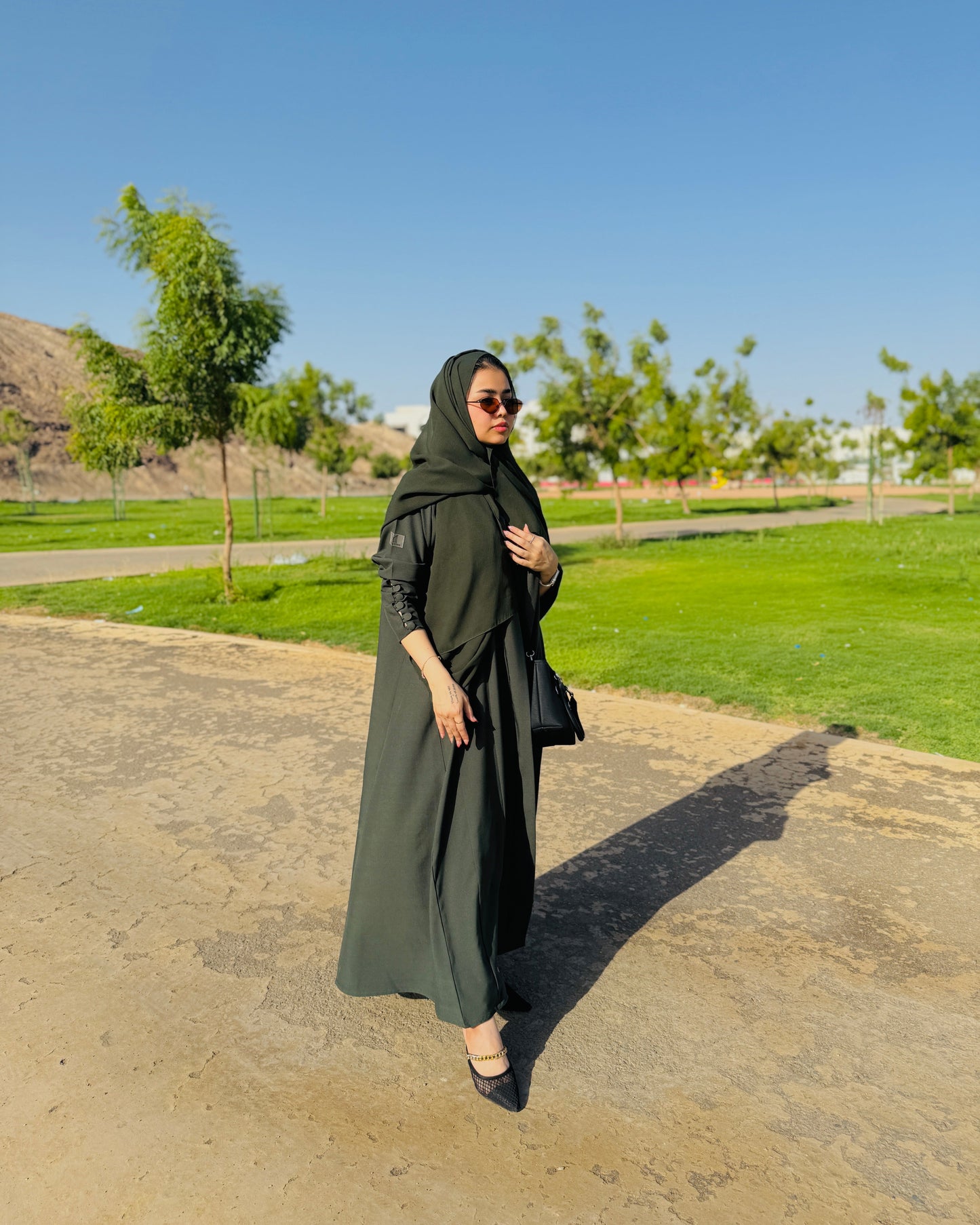 Woman in a green hijab and dress standing on a paved path with grass and trees in the background.