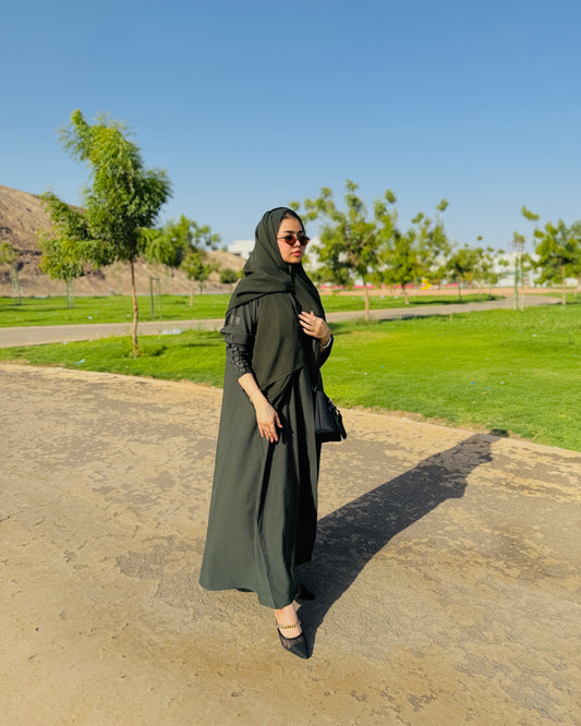 Woman in a green hijab and dress standing on a paved path with grass and trees in the background.